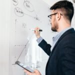 Close-up portrait of oung dark-haired man in glasses with laptop  writing a business plan on whiteboard. He wears blue shirt and dark jacket. View from side.
